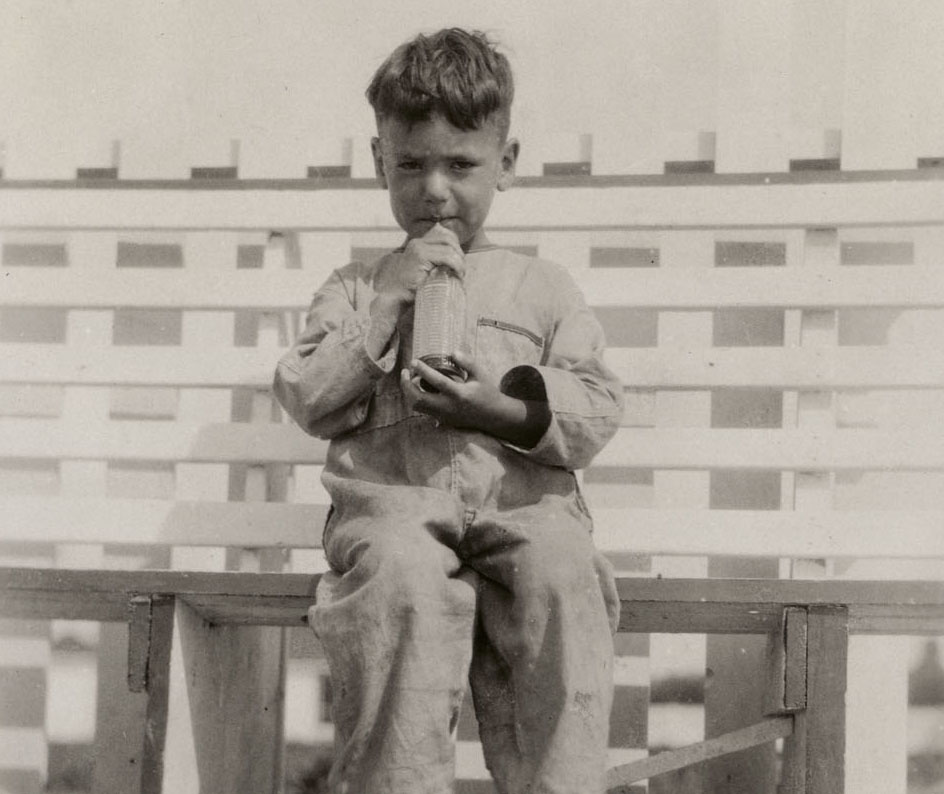 Black and white photograph of a young boy sitting on a bench drinking with a straw from a bottle. 
